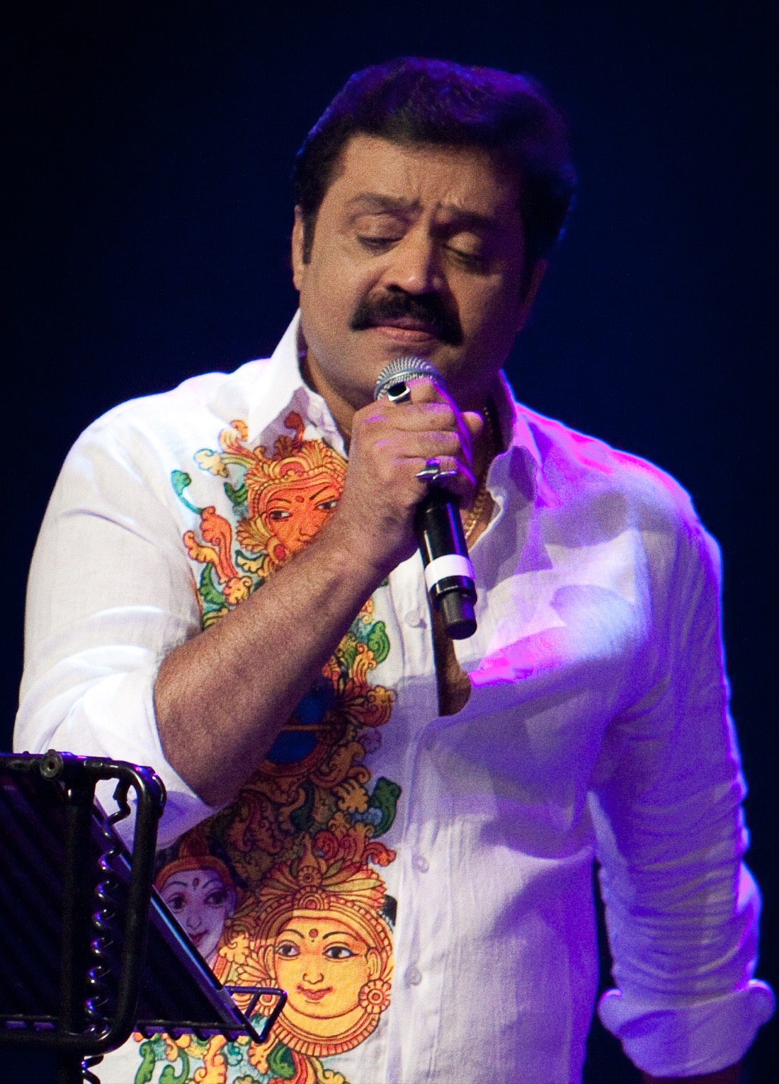 Suresh Gopi singing into a microphone on stage, wearing a shirt printed with Kerala mural figures