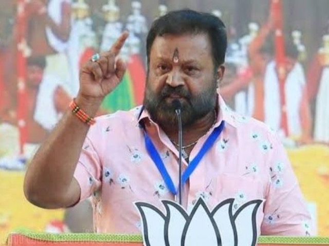 Suresh Gopi speaking from a lectern with one hand raised, in front of a backdrop showing a Kerala festival scene