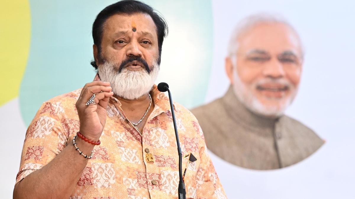 Suresh Gopi speaking into a microphone at a public event, gesturing with one hand, with a large poster of Prime Minister Narendra Modi behind him
